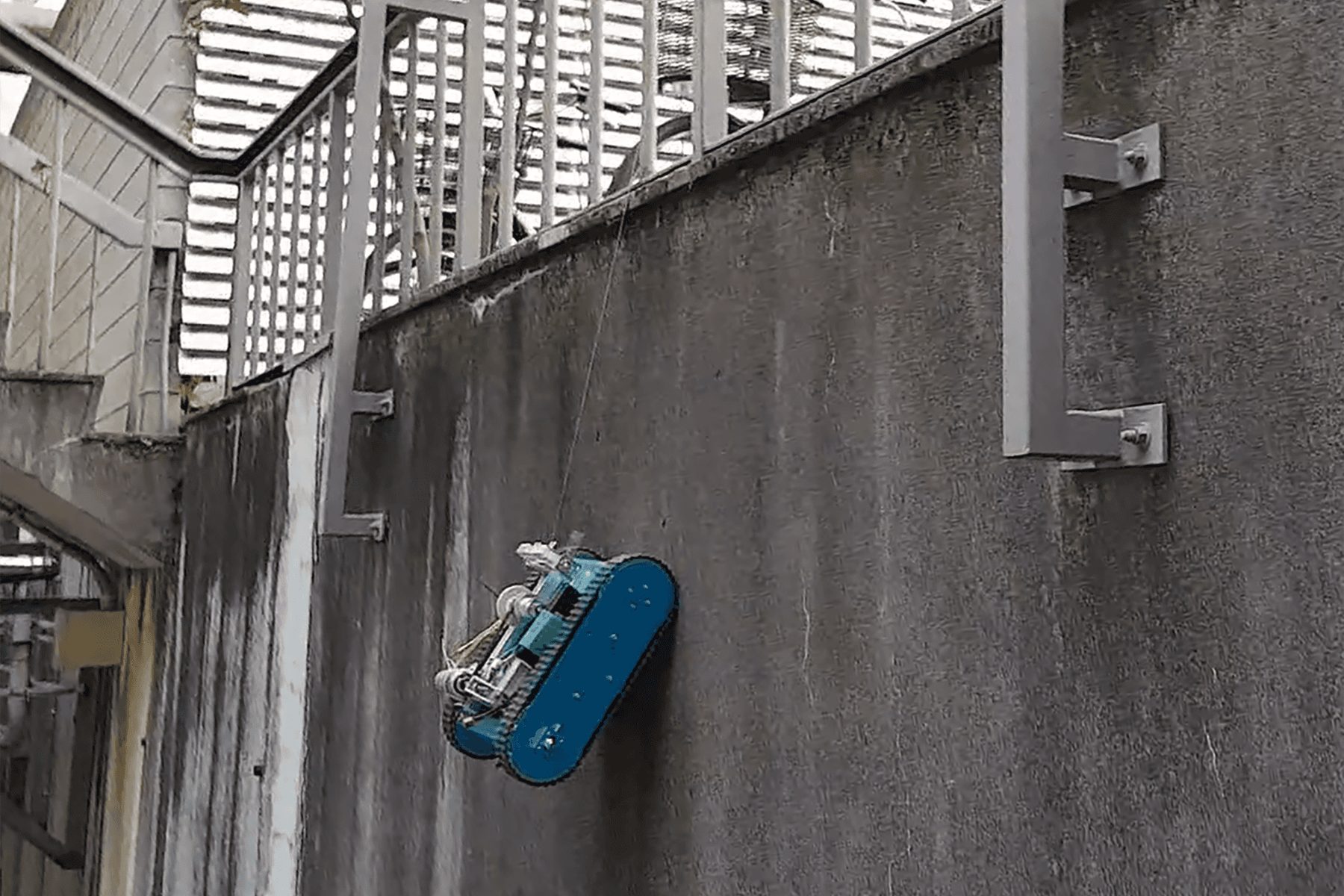 The tracked robot hanging on its tether halfway up a vertical concrete wall outdoors